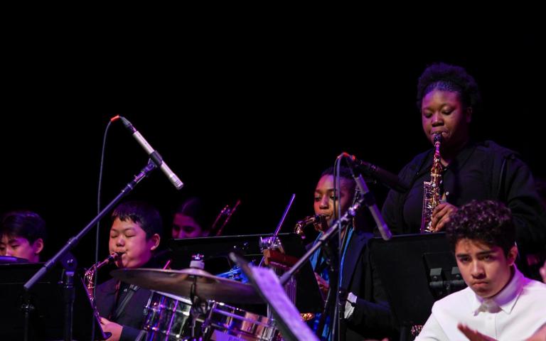 Six instrumentalists are pictured on the Berklee Performance Stage under a purple stage light. A young girl stands up while playing a soprano saxophone during her solo. 