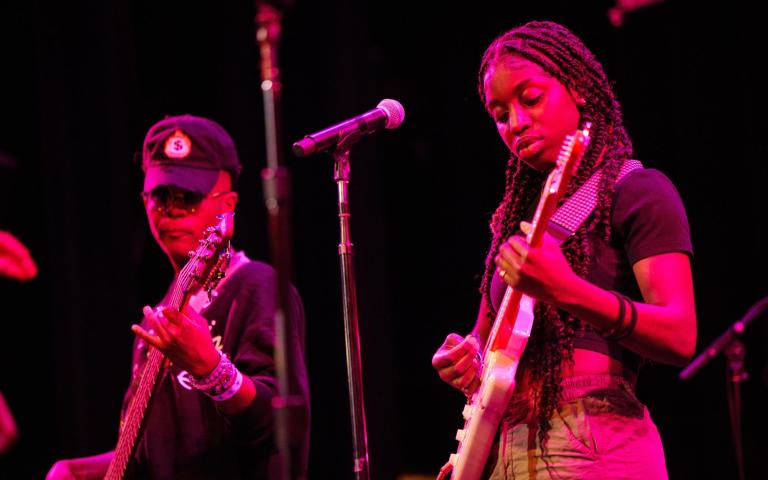 Two teenage performers stand on the Berklee Performance Center stage under a magenta stage light. The student on the left wears a black long-sleeved shirt and black cap while holding a dark-colored electric bass. The student on the right wears dark braids, a dark short-sleeved top and jeans while playing an electric guitar.