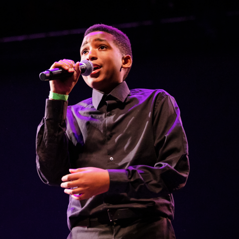 Pictured: Preparatory Academy student on-stage in a black button up and black pants, holding a microphone on-stage.
