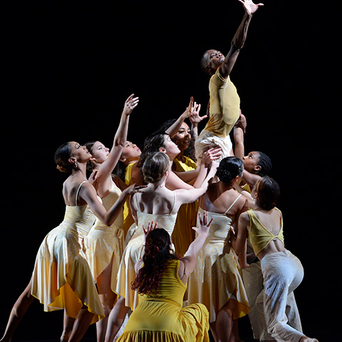 A group lifts one dancer who reaches toward the sky