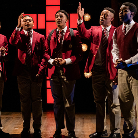 5 performers singing in matching outfits on stage: red jacket, black dress pants