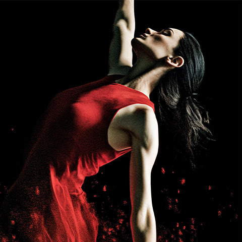 A girl with black hair and in a red dress is looking up and reaching her hand up in the air.