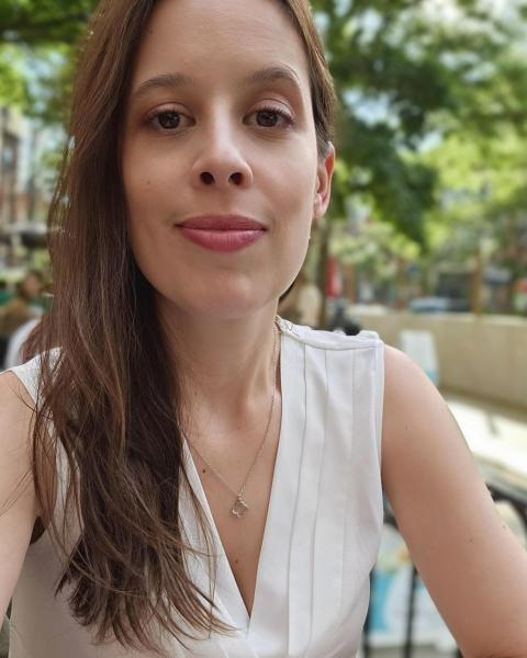 The photo portraits Dimayma McCauley, a White Latina in her late 30's with with long brown hair, wearing a white blouse and a delicate necklace, seated outdoors among blurred greenery and people.