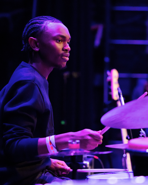 Student plays drum set wearing a black sweater and braids. 