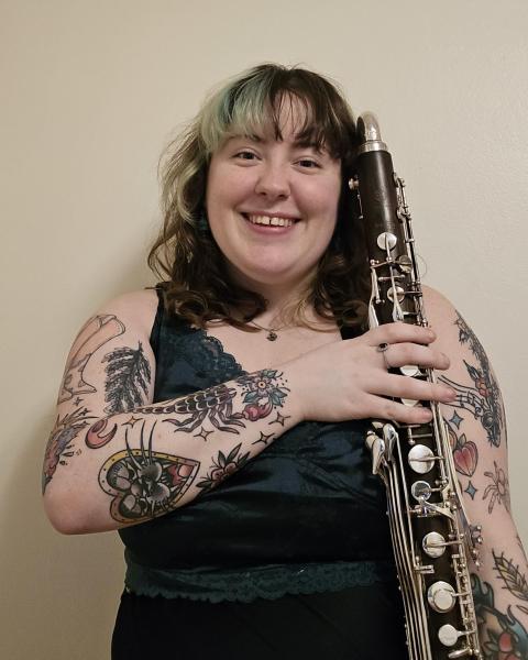 A picture of Tara Allen, Neurodiversity Program Manager. She is a white woman in her mid to late 20s, with shoulder length hair. The picture is of her torso up. She is holding her Bass Clarinet on the right, and smiling at the camera. Tara has green in her hair, and has two full tattoo sleeves. 