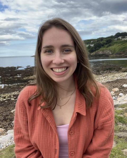 A smiling woman with light brown hair and an orange shirt sits in front of a coastal background.