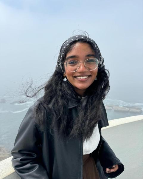 A smiling woman with glasses, dark brown hair, and a blue shirt stands in front of a coastal background.