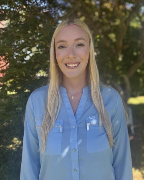Professional headshot of a woman with long blonde hair wearing a light blue shirt photographed in front of a tree with green leaves.