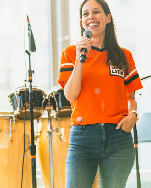 A medium shot of a woman with long, straight, dark brown&nbsp;hair and a&nbsp;light-to-medium complexion. She is captured mid-laugh while looking slightly upward and away from the camera, giving her an engaging, candid look. She is wearing a bright orange jersey-style T-shirt with black-and-white sleeve stripes, paired with blue jeans. In the background, musical instruments, including a microphone stand and conga drums, are visible in a brightly lit indoor space.