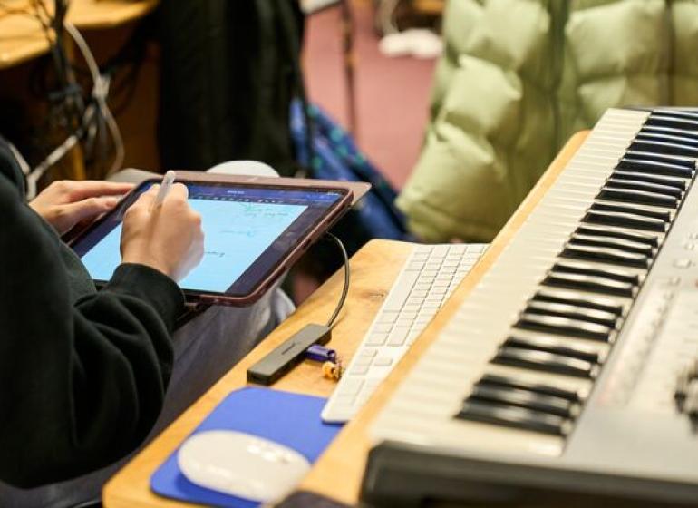 A person's hands writing on a digital tablet next to a MIDI keyboard workstation