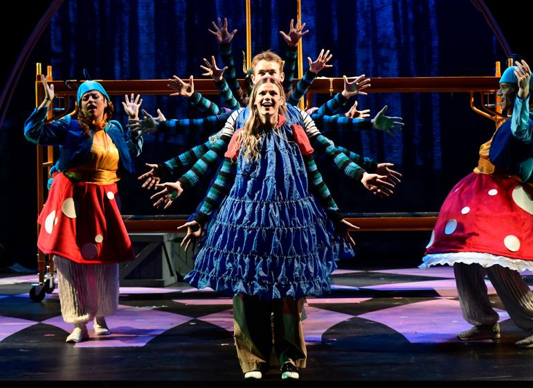 Theater students performing on stage in 'Alice by Heart' in a row with their arms spread out making a circle of arms