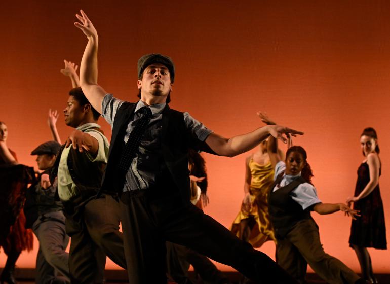 Dancers on stage in front of a n ornage background. Dancer in foreground has left hand lifted towards the ceiling with the right hands horizontal towards the right side.