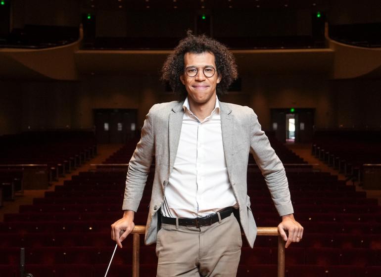 Jonathon Heyward smiling in a gray suit on a conducting podium in a concert hall.