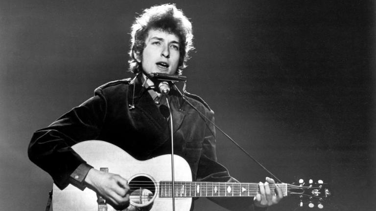 Black and white image of a young Bob Dylan performing on stage with an acoustic guitar and harmonica