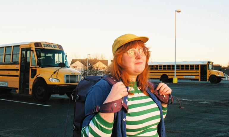 Singer-songwriter corook stands holding a backpack in a parking lot full of buses