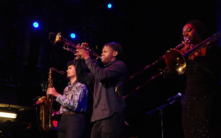Three high school-aged horn players stand on the Berklee Performance Stage - from left to right, a young man holds a tenor saxophone, another young man holds a trumpet, and a young woman holds a trombone. 