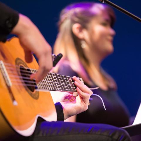 Closeup of a hand playing a nylon stringed guitar while a vocalist is in the background