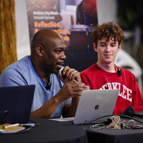 A bald-headed man with a blue t-shirt on holds a microphone facing an un-pictured audience, sitting next to a curly-haired student with a red sweatshirt on reading: "Berklee" -- both sit in front of laptops.