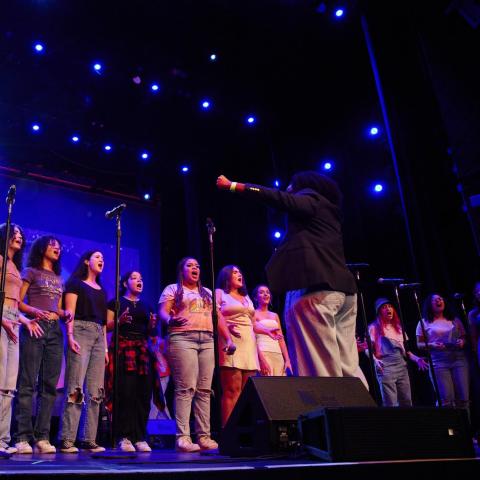 Fourteen young student vocalists stand in a semi-circle, looking towards their choir director while singing on the Berklee Performance Center stage - students wear a variation of jeans and light colored tops. A bright light blue light shines past a student's face.