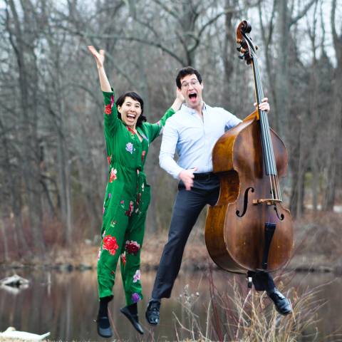 Two people jumping in the woods. One is wearing a green floral jumpsuit and one is in a button down shirt while holding a bass. 