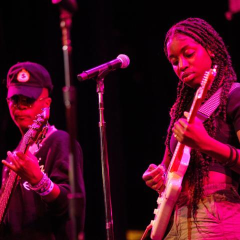 Two teenage performers stand on the Berklee Performance Center stage under a magenta stage light. The student on the left wears a black long-sleeved shirt and black cap while holding a dark-colored electric bass. The student on the right wears dark braids, a dark short-sleeved top and jeans while playing an electric guitar.