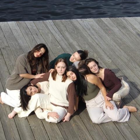 6 dancers resting against eachother on a pier by the water