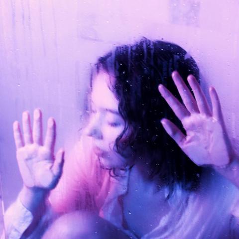 girl leaning up to glad or camera with both hands on the surface, monochromatic purple aura, head facing to the side, also leaning, glass is smudged. 