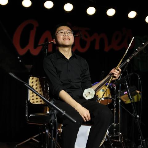 Yu sitting on stage holding a traditional string instrument, smiling under bright stage lights with drums in the background.