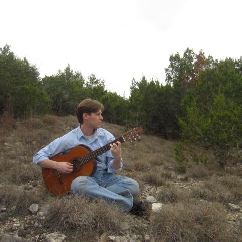 Jaret Cole sitting on a grassy hillside playing an acoustic guitar, surrounded by trees and looking off into the distance.