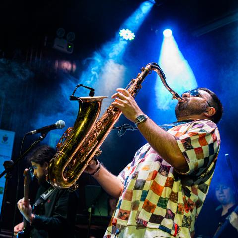 Diego Herrera playing a saxophone on stage under blue lights, with other musicians performing in the background.