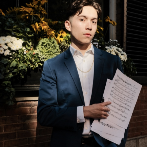 Montaño Quartet in a suit holding sheet music, standing outdoors in front of flowers and a window, looking directly at the camera.