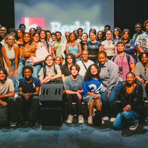 A large, diverse group of young adults smiles and poses together for a group photo on a theater stage. They are arranged in multiple rows, with some standing in the back and others sitting or kneeling on the stage floor in the front. Overhead stage lighting illuminates the group, and a projection screen in the background faintly displays the Berklee College of Music logo.