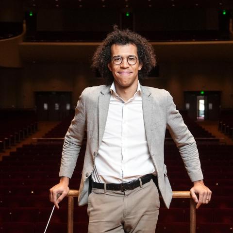 Jonathon Heyward smiling in a gray suit on a conducting podium in a concert hall.