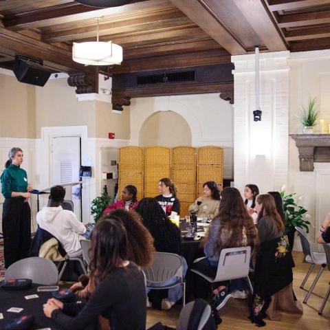An indoor group presentation taking place in a room featuring coffered wooden ceilings and a large fireplace decorated with a clock and small plants. On the left, a female-presenting speaker wearing a teal turtleneck and black trousers stands facing the room, addressing an audience seated at round tables with dark tablecloths.
