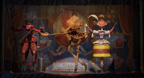 A film still of a wooden Pinocchio on stage in between a devil puppet to the left and a woman puppet to the right