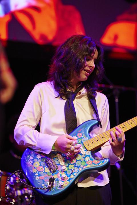 A High School Academy guitarist/student in a white button up shirt, black tie, and long brown hair holding a metallic blue guitar with sticker decorations.