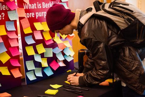 A student writes on a post-it; a poster board filled with post-its is in the background