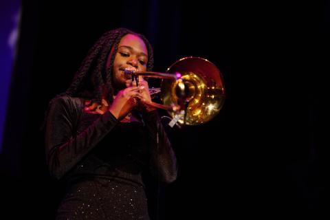 A young girl with dark braids, wearing a black dress plays a large gold trombone on-stage.