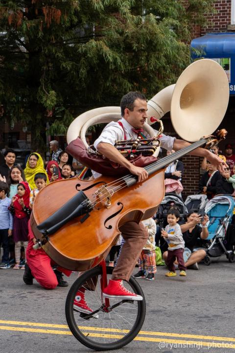 Colescott Rubin riding a unicycle while holding an acoustic bass and a tuba during a parade