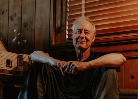 Bruce Hornsby smiles, sitting on the floor by a window with the shades drawn