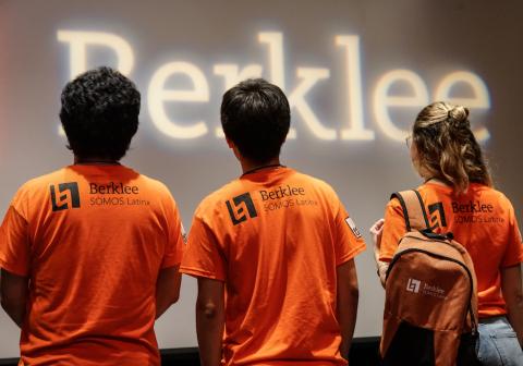 Three individuals, two young men and one young woman in orange shirts pose in front of a screen displaying the word "Berklee".