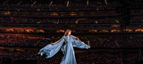 Taylor Swift in a flowy dress on stage with a backdrop of a packed stadium of fans