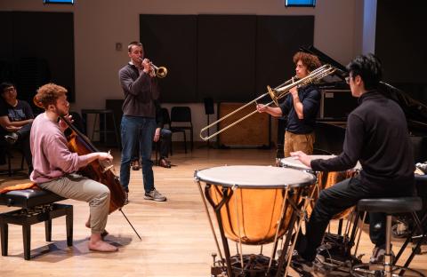 Four musicians playing together, a cellist, a timpanist, a trombonist, and a trumpet player.