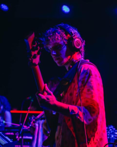 Chase Behar wearing headphones and a patterned shirt plays electric bass on stage under dramatic blue and pink lighting, focused intently on the instrument.