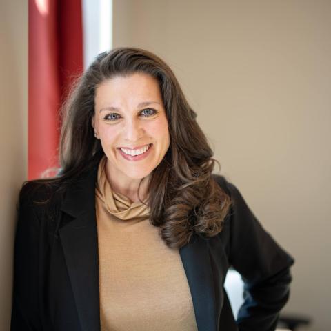 Woman with brown hair wearing black blazer and gold top, leaning against a wall, smiling