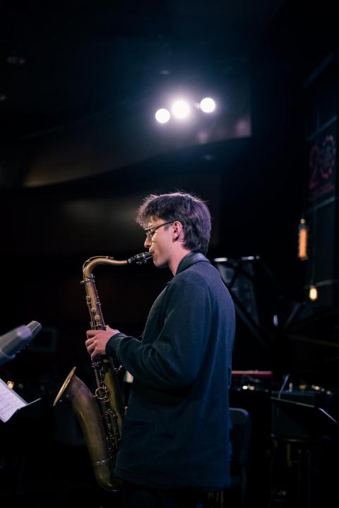 Boy with glasses from side angle playing the saxophone under stage lights