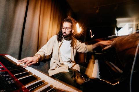 Photo of Yessaï Karapetian playing a piano and keyboard with each hand. He has long curly black hair, a beard, and glasses.