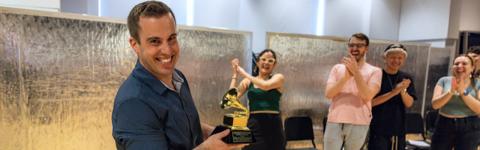Stephen Spinelli looking at the camera and holding his Grammy trophy while students in the background smile and clap