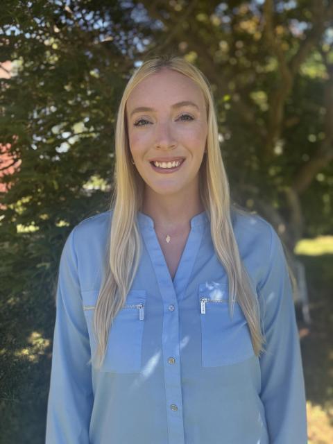 Professional headshot of a woman with long blonde hair wearing a light blue shirt photographed in front of a tree with green leaves.