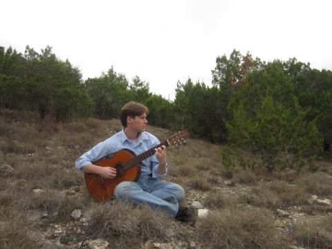 Jaret Cole sitting on a grassy hillside playing an acoustic guitar, surrounded by trees and looking off into the distance.